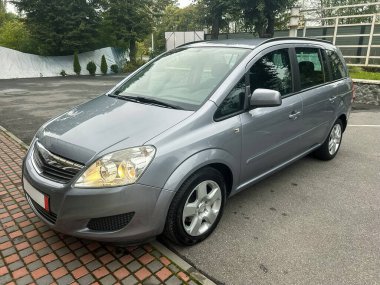 Gümüş renkli bir minivan, yemyeşil ve açık renkli bulutlarla çerçevelenmiş düz bir yolun kenarında duruyor ve sabahın erken saatlerinde sakin ve sakin bir atmosfer yaratıyor..