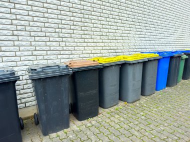 Çeşitli renklerde bir dizi geri dönüşüm kutusu desenli tuğla bir duvara karşı güzelce oturur. Bu sahne, şehirde uygun atık imhası ve çevresel bakımı teşvik etmektedir..