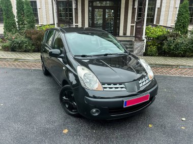 Zarif mimarisi olan çağdaş bir yapının yanına parlak siyah bir araba park edilmiştir. Sahnede bulutlu bir gökyüzü ve canlı bir yeşillik var, atmosferi güçlendiriyor..