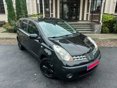 Parlak siyah bir hatchback, yemyeşil bitkiler ve açık gökyüzü tarafından çerçevelenmiş güzel bir tuğla binanın önünde zarif bir şekilde duruyor. Arabanın parlak yüzeyi, bakımını iyi yaptığını gösteriyor..