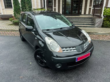 Gösterişli siyah bir araba, gür ağaçlar ve çağdaş bina özellikleriyle çerçevelenmiş şık bir garaj yolunda durur. Sahne bulutlu bir günde sakin bir atmosfer yakalar..