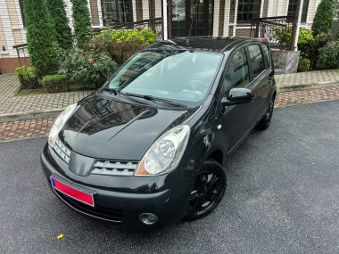 Pürüzsüz bir asfalt zemine park etmiş parlak siyah bir hatchback. Araba yemyeşil ve modern binalarla çevrili. Huzurlu bir atmosfer yaratıyor..