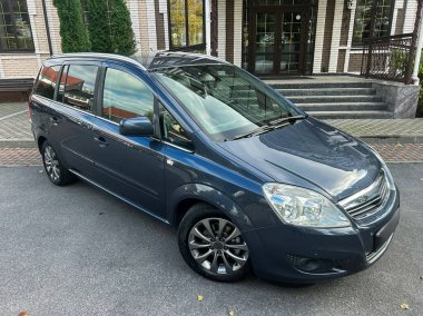 Bu mavi minivan geniş pencereli çağdaş bir binanın önünde zarifçe park edilmiştir. Sahne güneş ışığı ile aydınlanır ve yemyeşil bir çerçeve ile çerçevelenir, sakin bir atmosfer yaratır..