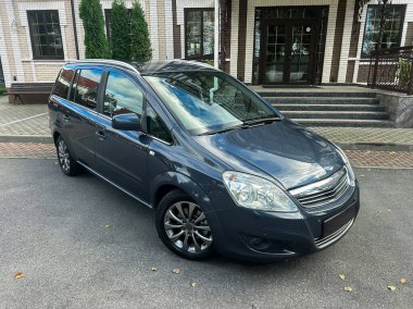 Şık bir minivan düzgün bir şekilde sofistike bir binanın önüne park edilmiştir. Dekor yeşil ve parlak güneş ışığına sahip. Aracın parlak dış görünüşünü gösteriyor..