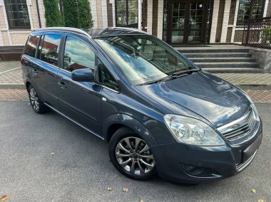 Modern bir binanın önüne park edilmiş şık bir aile arabası, parlak yüzeyine yansıyan ışığı yansıtıyor. Güneş yerde hafif gölgeler bırakıyor..