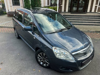 Modern bir yapının yakınındaki pürüzsüz bir yüzeye park edilmiş şık gri bir minivan. Parlak gökyüzü aracı yansıtıyor, sakin bir atmosfer yaratıyor..