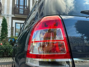 Gösterişli siyah bir araba büyüleyici bir bahçenin yanına park edilmiş, parlak arka lambası sergileniyor. Güneş ışığı lensten yansıyarak huzurlu ortamda sıcak bir atmosfer yaratır..