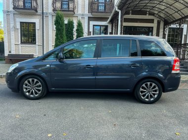 Gri bir minivan temiz bir caddede duruyor. Etrafı yeşilliklerle çevrili ve güneşli bir günün sıcaklığının tadını çıkarıyor..