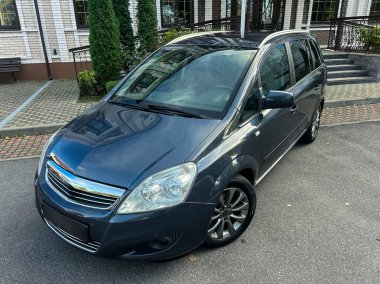 Parlak mavi minivan zarif bir şekilde güzel bir garaj yolunda duruyor, yemyeşil ve bakımlı bir evle çevrili, huzurlu bir öğleden sonra atmosferini yansıtıyor..