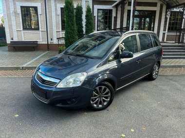 Gösterişli gri bir minivan şık bir çağdaş evin önüne düzgün bir şekilde park edilmiştir. Güneş ışıl ışıl parlıyor, aracın temiz çizgilerini ve bahçesini aydınlatıyor.