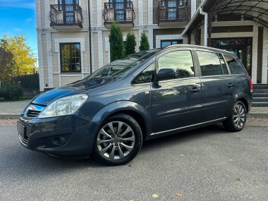 Modern gri bir minivan, güzel bir konutun yanındaki pürüzsüz bir garaj yolunda, ağaçlar rüzgarda hafifçe sallanıyor ve güneş tepemizde parıldıyor..