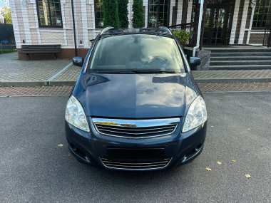 Modern bir mimari yapının önünde mavi bir araç duruyor. Güneş ışığı parlak yüzeyinden yansıyarak sakin ve davetkar bir sahne yaratır..