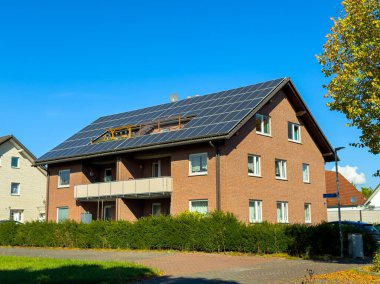 Çatıda güneş panelleri olan güzel bir ev güneşli bir sonbahar öğleden sonrasında, yemyeşil çalılar ve açık mavi bir gökyüzü ile parıldıyor..