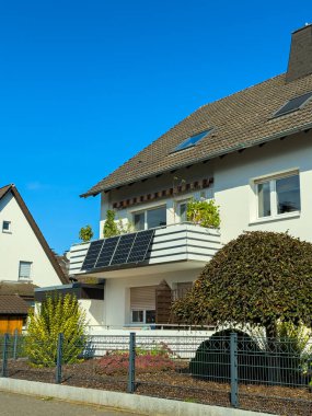 Güneşli bir günde, sakin bir mahallede geçen, bitkiler ve güneş panelleriyle süslenmiş bir balkonu olan çağdaş bir ev vurgulanıyor. Çimenli bir ağaç sahneye cazibe katar..