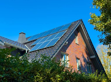 Güneş ışığı, kırsal bir tuğla evin eğimli çatısına kurulmuş güneş panellerine parlıyor. Bereketli yeşillik, parlak mavi bir gökyüzünün altında çevreci yaşamı gözler önüne seriyor..