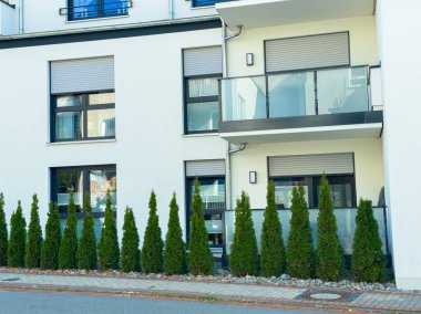 Beyaz duvarları olan çağdaş bir binada, önünde yüksek yeşil ağaçlar sergilenerek, yürüyüş yolunun yakınında sakin bir atmosfer yaratılıyor. Zarif zarafet bu kentsel yaşam alanını vurgular.