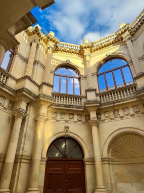 Görkemli bir avlu, uzun pencereleri, zarif pervazları ve yukarıdaki parlak mavi gökyüzü ile süslenmiş bir mimariyi gözler önüne seriyor. Tarihin ve zanaatkarlığın zamansız bir duygusunu yansıtıyor..