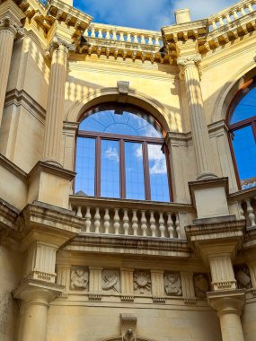 Sıcak güneş ışığı, eski bir binanın zarif kemerlerini ve detaylı taş işçiliğini aydınlatır. Kabarık bulutlu, parlak mavi gökyüzüne karşı çarpıcı bir kontrastlık yaratır..