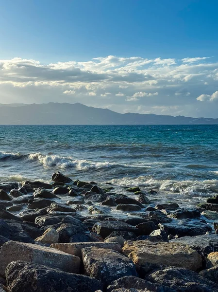 Güneş batarken, yumuşak dalgalar kayalık bir kıyıya vurur ve suyun üzerine sıcak bir ışık yayar. Uzak dağlar huzur verici bir manzara yaratır, deniz tarafından dinlenmeye ve yansımaya davet edilir..