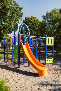 Parlak renkli oyun alanı, güneşli bir parkta durur. Eğlenceli bir turuncu kaydırak, tırmanma çerçeveleri ve kumlu bir taban vardır. Çocukların keşfetmesi ve dışarıda vakit geçirmesi için mükemmeldir..