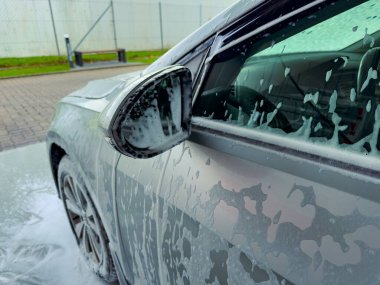 Parlak gümüş renkli araç, gün ışığında, yoğun köpükler ve köpüklerle kaplanarak, düzenli bir oto yıkamada yıkanırken, kenar mahallelerden akan sular parıldar..