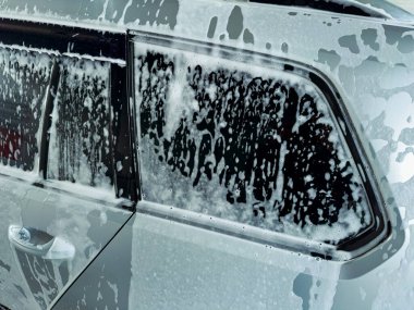 Parlak güneş ışığı yeni yıkanmış bir arabanın yüzeyinde parıldıyor, kalın köpüklü sabunla kaplanmış. Baloncuklar eğlenceli bir doku oluştururken temizlik süreci titiz bir şekilde işliyor..