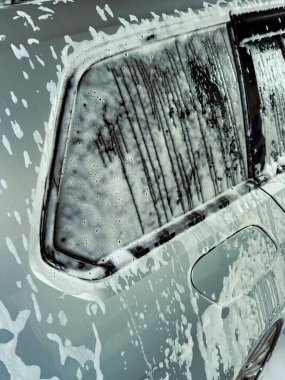 Sabun köpükleri parlak, gümüş renkli bir arabayı kaplıyor. Suyun yanından aktığı neşeli bir sahne yaratıyor. Güneşli bir ortamda araba temizlemenin canlı bir öğleden sonrasını sergiliyor..