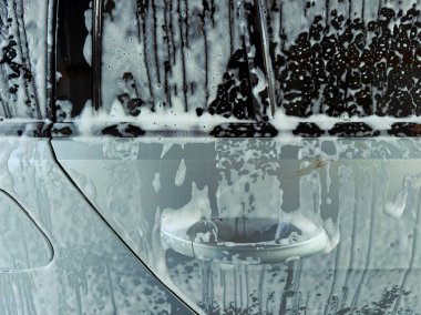 Köpüklü sabun arabanın kenarını kaplıyor, parlak güneş altında parlarken su damlıyor, taze ve temiz bir görünüm yaratıyor. Araç bakımı için mükemmel bir yer..