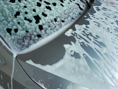 Köpüklü kabarcıklar gümüş bir arabanın parlak yüzeyini kaplarken, güneş ışığı parlak boyayı yansıtıyor ve hafta sonu temizlik ritüelinin neşesini vurguluyor..