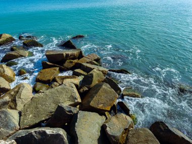 Parlak turkuaz dalgalar huzurlu bir sahil şeridinde engebeli kayalara hafifçe çarpar, açık mavi gökyüzünün altında deniz kenarında sakin bir günün özünü yakalar..