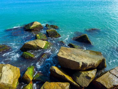 Güzel bir sahne, turkuaz suların kıyı şeridi boyunca yavaşça süzülen kayaları, sakin ve sakin bir atmosfer yaratarak berrak mavi gökyüzünün altında.