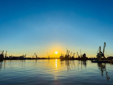 Güneş ufukta batarken, yoğun bir limanın üstündeki gökyüzünü canlı bir turuncu ışıltı kaplıyor. Turnalar ve gemiler dik durup sakin sulara yansıyan pitoresk bir ufuk çizgisi oluştururlar..