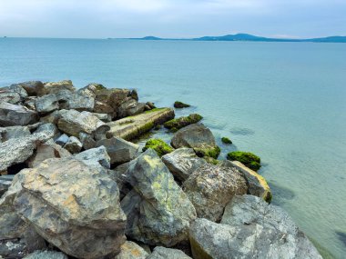Dalgalar, geniş kayaların doğal heykeller yarattığı engebeli bir kıyı şeridine nazikçe çarpar. Uzak tepeler, yumuşak bir gökyüzünün altında sakin bir okyanusa karşı yükseliyor. Huzur ve yalnızlığı çağrıştırıyor..