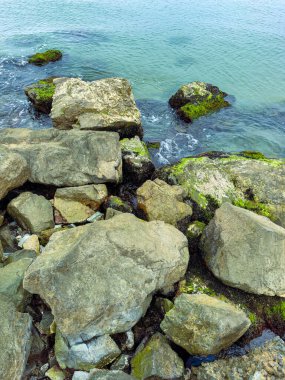 Büyük, yıpranmış kayalar, deniz yosunlarıyla kaplı kıyılar, güneşli bir günde sakin turkuaz suyla nazikçe kucaklanarak, doğanın güzelliğinin sakin bir manzarasını yaratıyor..