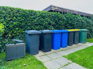 Bulutlu bir öğleden sonra, sakin bir mahallede çevre dostu uygulamaları temsil eden yemyeşil bir çitin yanında, çeşitli renklerdeki çöp kutuları düzgün bir şekilde duruyor..