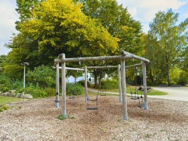 Basit bir oyun parkında, büyük ağaçların gölgesinde kurulu ahşap salıncaklar çocukların eğlenmesi için huzurlu bir yer sağlıyor. Yumuşak zemin ahşap parçalarıyla kaplı..