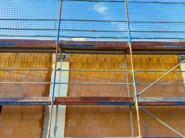 İşçiler, açık mavi gökyüzüne karşı sağlam bir iskeleyle bina bakımına hazırlanıyorlar. Duvarda devam eden inşaat çalışmalarını gösteriyorlar..