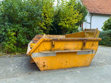 Parlak sarı bir çöp bidonu, sakin bir mahallede ağaçlar ve çalılarla çevrili asfalt bir yolda belirgin bir şekilde duruyor. Rengi yeşillikle ve yakınlardaki evlerle çelişiyor..