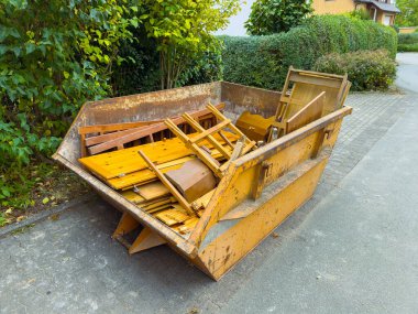 Parlak sarı bir çöp konteyneri, huzurlu bir cadde boyunca park etmiş ahşap kırıntılar ve molozlarla dolup taşıyor. Yeşil yapraklar, bulutlu bir günde banliyö atmosferini yükselterek olay yerini çerçeveler..