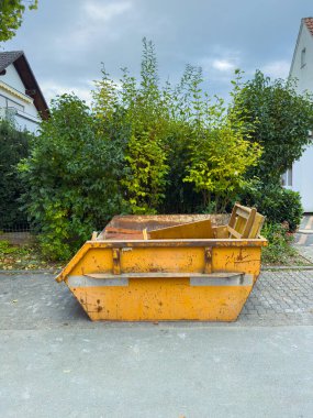 Canlı yeşil çalılarla çevrili, huzurlu bir banliyö yolu boyunca parlak sarı bir çöp konteynırı park edilmiştir. Bulutlu gökyüzü sahneye karamsar bir atmosfer ekler..