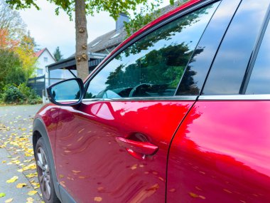 Parlak kırmızı bir araba, düşen yapraklarla dolu sakin bir caddenin yanında duruyor. Aracın çevresindeki sonbahar renkleri huzurlu bir öğleden sonra atmosferini yansıtıyor ve gösterişli tasarımını sergiliyor..