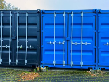 İki nakliye konteynırı, biri mavi ve biri siyah, bir depoda yan yana durur, kaldırım taşlarıyla çevrili ve yapraklarla kaplı, gri bir gökyüzünün altında, tipik bir sonbahar günü..