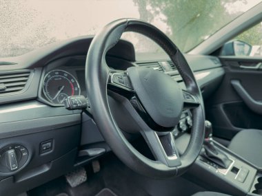 Şık bir arabanın içinde, siyah bir direksiyon parlar, gelişmiş sürüş için kontrolleri gösterir. Yağmur rahat bir atmosfer yaratır, dışarıdaki manzarayı bulanıklaştırır..