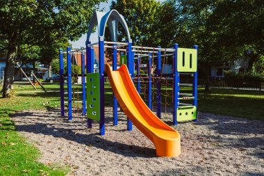 Parlak renkli oyun alanı yapısı yemyeşil bir parkta duruyor, uzun bir kaydırak ve tırmanma çubukları var, neşeli çocukların sıcak ve güneşli bir günde eğlenmesine hazır..