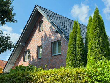 Güneş ışığı sıcacık bir evin güzel tuğla cephesini yıkıyor. Orada uzun yeşil ağaçlar ve canlı çalılar huzurlu bir atmosfer yaratıyor. Sahne sıcaklığı ve sükuneti yakalıyor..