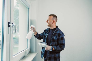 Camları sprey şişeyle spreylemekten zevk alan bir adam. Güneş ışığı odayı dolduruyor, ferahlatıcı bir atmosfer yaratıyor, o da bu arada mekanı parlatmaya odaklanıyor..