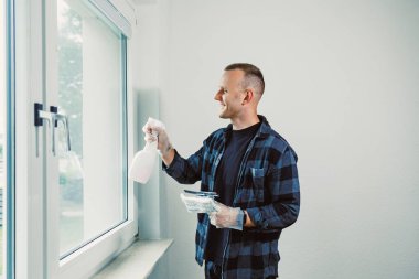 Damalı gömlekli bir adam, çağdaş bir dairede pencereyi temizlerken gülümser ve doğal ışıkla dolu yeni bir atmosfer yaratır..