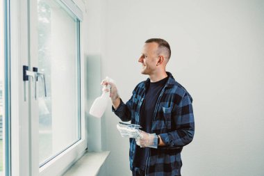 Neşeli genç bir adam güneşli bir odada pencereleri temizliyor, elinde bir sprey şişesi ve bez tutuyor, görevinden memnuniyet ve neşe duyuyor..