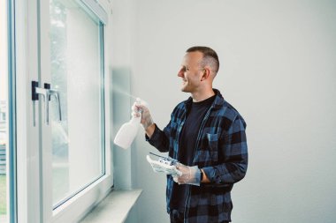 Yüzünde bir gülümsemeyle, bir adam cam camına temizleyici sıkar, parlak güneş ışığı temiz, dağınık odaya akar ve temizlik görevinden zevk alır..