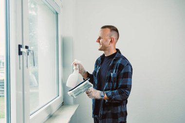 Kareli gömlekli neşeli bir adam sprey bir şişe ve cam temizleme aleti tutuyor. Parlak bir odada duruyor, güneş ışığında camı parlatmaya hazır..